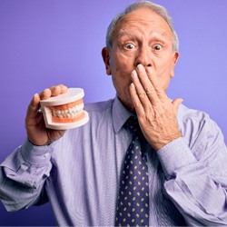 man in front of a purple background covering his mouth and holding dentures 